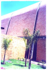 Vista externa de edifício com cobertura abobadada revestida em tijolos, parede de alvenaria e jardim com palmeiras em primeiro plano — P0131 — SESC - Piracicaba