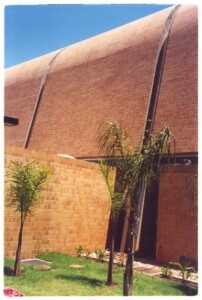 Vista externa de edifício com cobertura abobadada revestida em tijolos, parede de alvenaria e jardim com palmeiras em primeiro plano — P0131 — SESC - Piracicaba