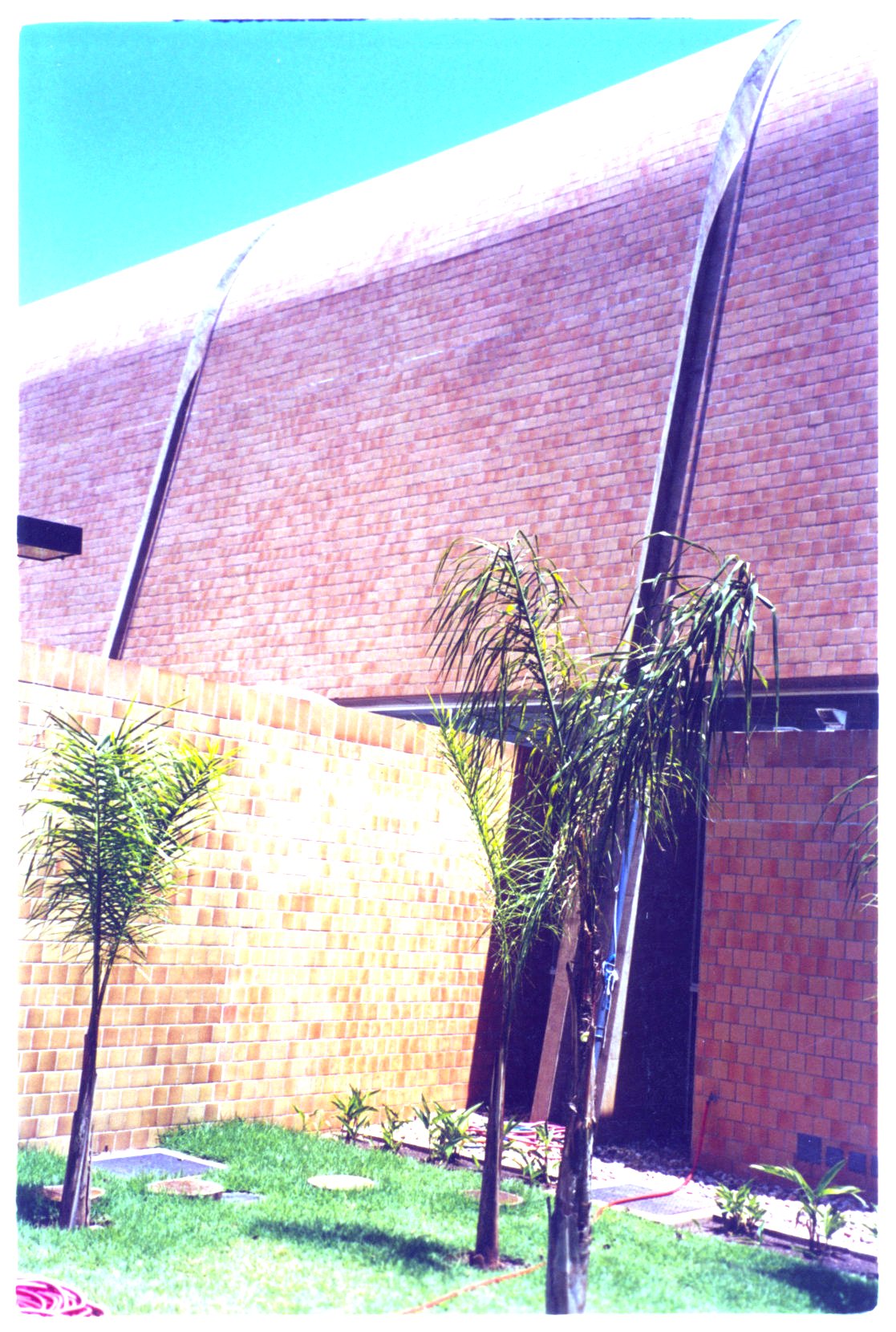 Vista externa de edifício com cobertura abobadada revestida em tijolos, parede de alvenaria e jardim com palmeiras em primeiro plano — P0131 — SESC - Piracicaba — F009-P0131-0000-S03-D00020