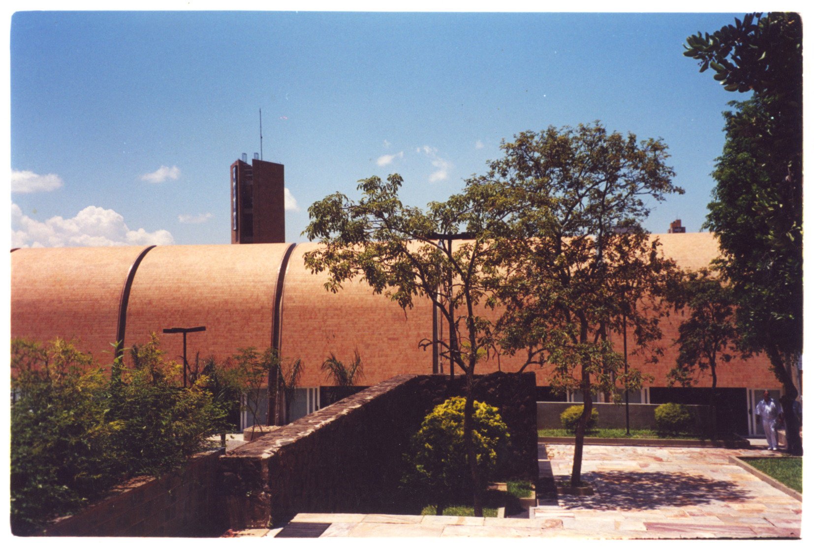 Vista externa de edifício com cobertura curva em tijolo aparente e torre ao fundo, a partir de jardim com muro de pedra — P0131 — SESC - Piracicaba — F009-P0131-0000-S03-D00017