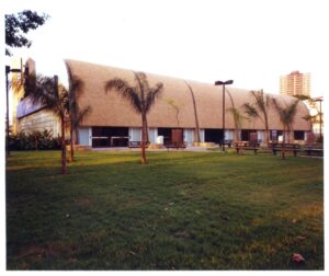 Vista externa de edifício com fachada curva, cercado por gramado, árvores e postes de iluminação — P0131 — SESC - Piracicaba