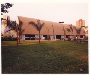 Vista externa de edifício com fachada curva, cercado por gramado, árvores e postes de iluminação — P0131 — SESC - Piracicaba