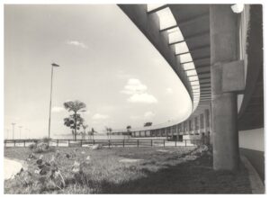 Vista externa de edifício com fachada curva em concreto aparente, marquise com brises e grande área gramada em primeiro plano — P0015 — SESI — Ribeirão Preto