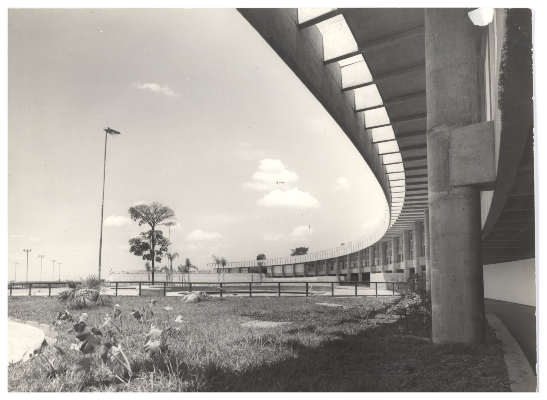 Vista externa de edifício com fachada curva em concreto aparente, marquise com brises e grande área gramada em primeiro plano — P0015 — SESI — Ribeirão Preto — F009-P0015-1973-S03-D00011
