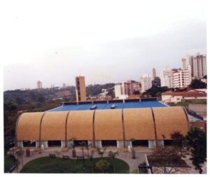 Vista externa de edifício com fachada curva em painéis terracota, elevado sobre pilotis, com praça e vegetação ao redor — P0131 — SESC - Piracicaba