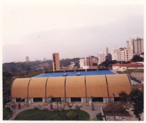 Vista externa de edifício com fachada curva em painéis terracota, elevado sobre pilotis, com praça e vegetação ao redor — P0131 — SESC - Piracicaba