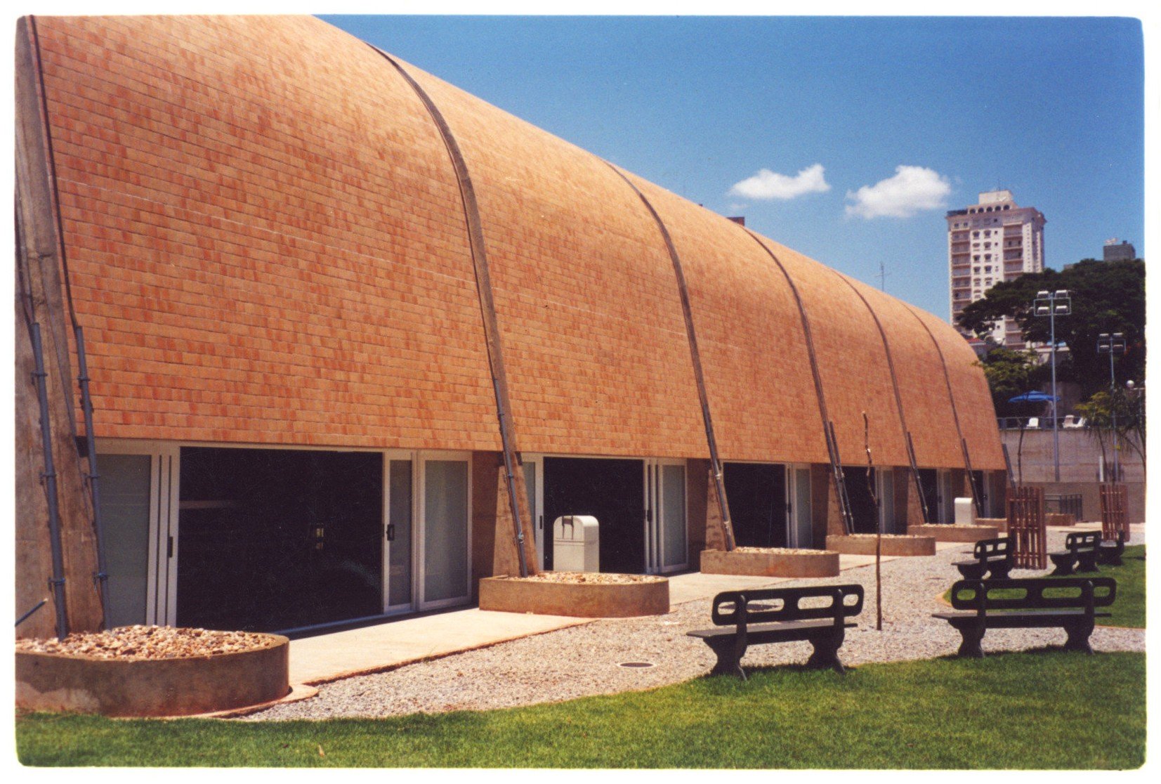 Vista externa de edifício com fachada revestida em telhas cerâmicas e aberturas envidraçadas, com mobiliário de piquenique na área externa (imagem invertida) — P0131 — SESC - Piracicaba — F009-P0131-0000-S03-D00016