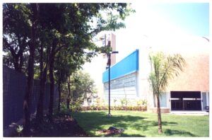 Vista externa de edifício com painel azul e pátio de pedra, com árvores em primeiro plano — P0131 — SESC - Piracicaba