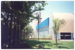 Vista externa de edifício com painel azul e pátio de pedra, com árvores em primeiro plano — P0131 — SESC - Piracicaba