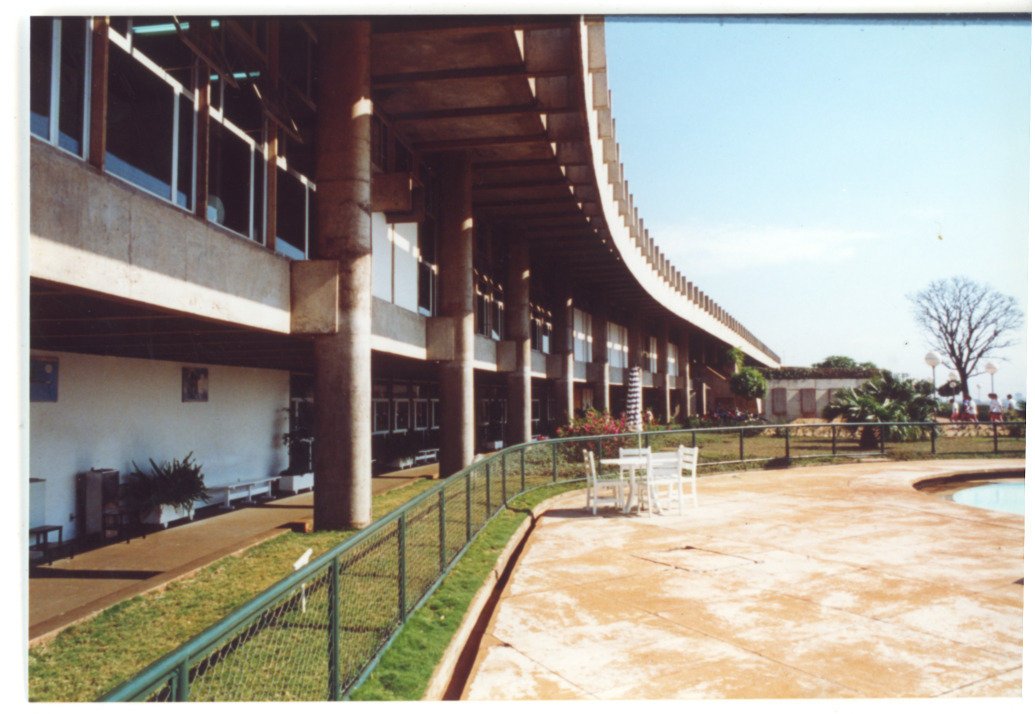 Vista externa de edifício de linhas curvas sustentado por pilotis, com varanda de piso superior, grade metálica delimitando gramado, mesa e cadeiras próximas à piscina, e árvores ao fundo — P0015 — SESI — Ribeirão Preto — F009-P0015-1973-S03-D00054