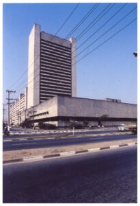 Vista externa de edifício em concreto com torre vertical de brises horizontais sobre embasamento alongado, a partir da avenida — P0018 — Correios — Sede Regional e Centro de Triagem