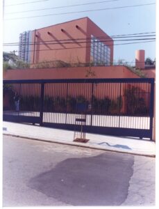 Vista externa de edifício em tom terracota, com grade metálica, árvores e calçada em primeiro plano — P0131 — SESC - Piracicaba