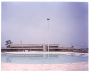 Vista externa de edifício institucional, com placa indicando "SESI", piso pavimentado e poste de iluminação (foto registrada invertida) — P0055 — SESI — São José dos Campos