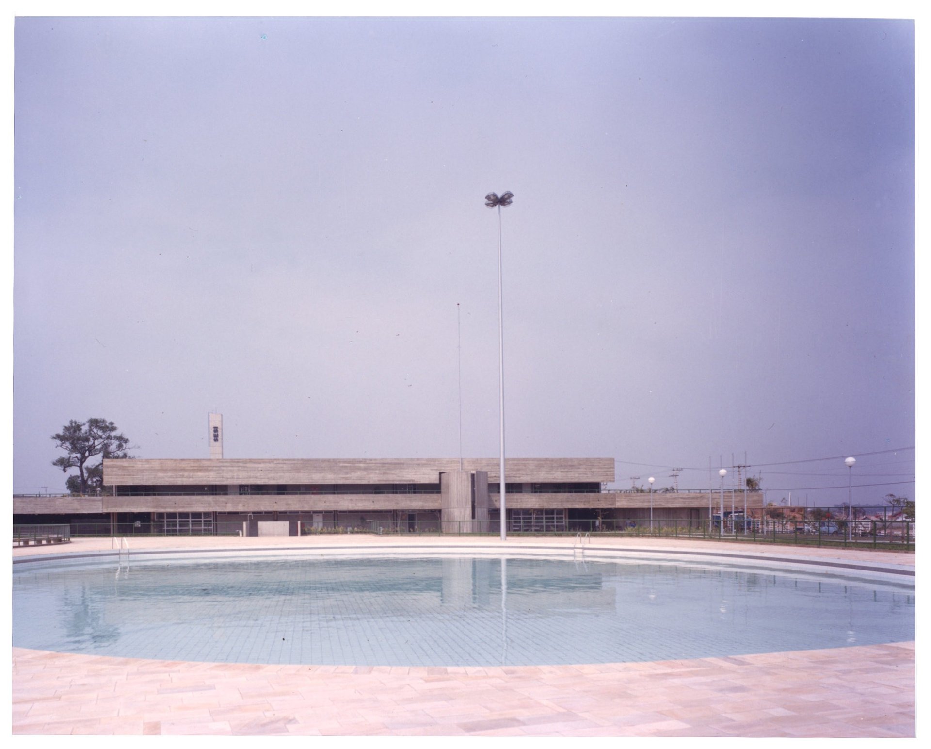 Vista externa de edifício institucional, com placa indicando "SESI", piso pavimentado e poste de iluminação (foto registrada invertida) — P0055 — SESI — São José dos Campos — F009-P0055-1979-S03-D00011
