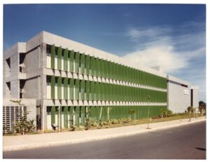 Vista externa de edifício modernista com brises horizontais verdes e cobertura em treliça, em dia de céu claro — P0060 — SESI — Marília