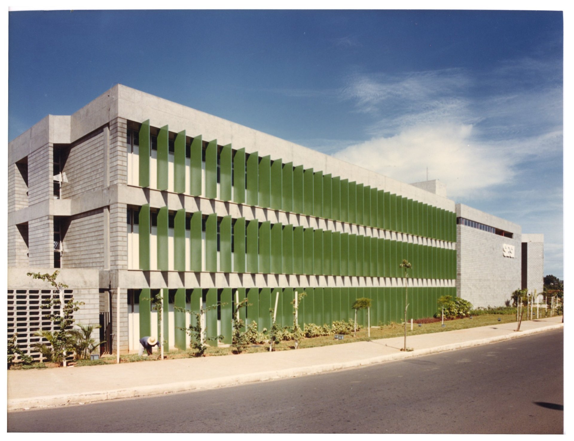 Vista externa de edifício modernista com brises horizontais verdes e cobertura em treliça, em dia de céu claro — P0060 — SESI — Marília — F009-P0060-1984-S03-D00021