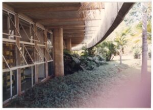 Vista externa de edifício sobre pilotis, com fachada de esquadrias de vidro basculantes e jardim com vegetação tropical ao longo do passeio coberto — P0015 — SESI — Ribeirão Preto