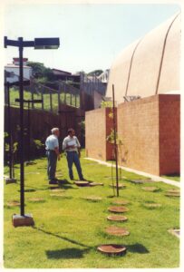 Vista externa de jardim com piso de discos de madeira sobre grama, postes de iluminação e muro de tijolos aparentes, ao lado de edifício com cobertura abobadada; duas pessoas conversam em primeiro plano — P0131 — SESC - Piracicaba