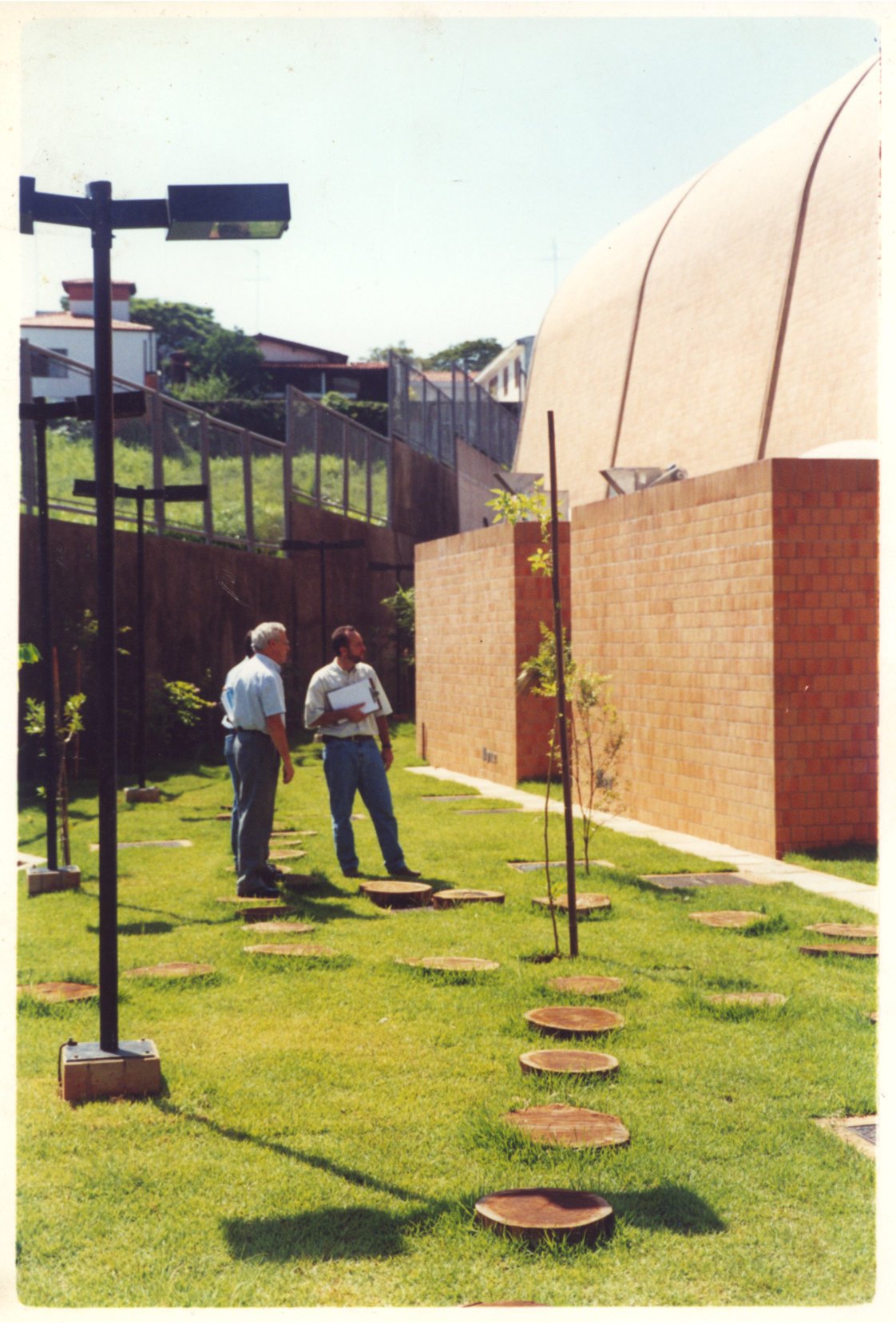 Vista externa de jardim com piso de discos de madeira sobre grama, postes de iluminação e muro de tijolos aparentes, ao lado de edifício com cobertura abobadada; duas pessoas conversam em primeiro plano — P0131 — SESC - Piracicaba — F009-P0131-0000-S03-D00019