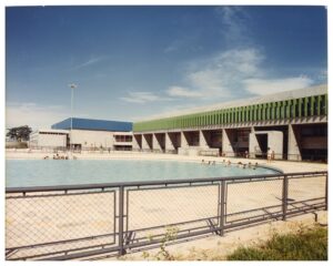 Vista externa de piscina pública com banhistas, em primeiro plano cercada por grade metálica, tendo ao fundo edifício de concreto com elementos verticais verdes e cobertura azul — P0060 — SESI — Marília