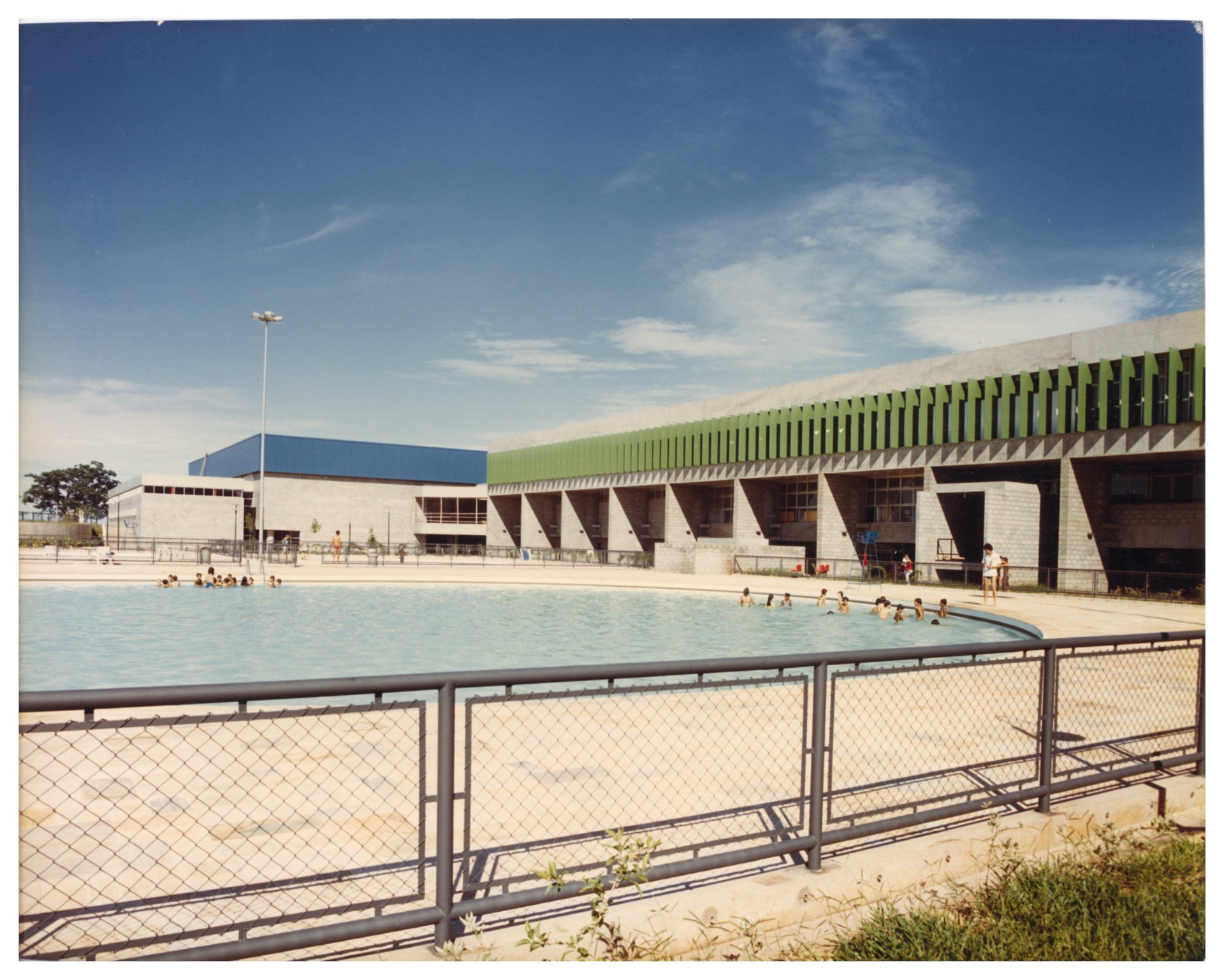 Vista externa de piscina pública com banhistas, em primeiro plano cercada por grade metálica, tendo ao fundo edifício de concreto com elementos verticais verdes e cobertura azul — P0060 — SESI — Marília — F009-P0060-1984-S03-D00023