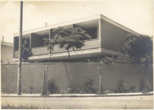 Vista externa de residência modernista, com pavimento superior em balanço sobre pilotis, muro de fechamento frontal e vegetação ao redor — P0001 — Residência Aecio Aeouche de Toledo