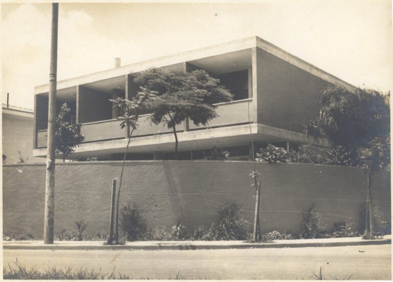 Vista externa de residência modernista, com pavimento superior em balanço sobre pilotis, muro de fechamento frontal e vegetação ao redor