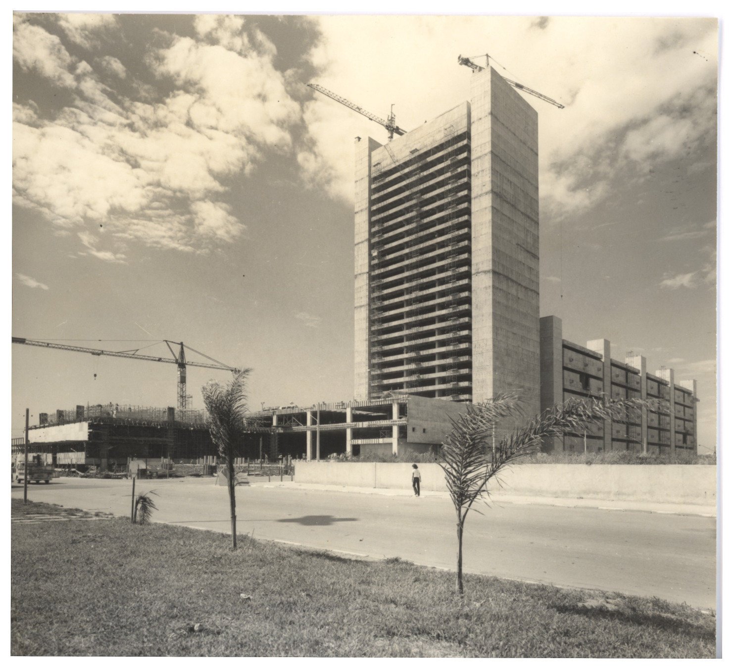 Vista externa de torre em construção com guindastes, ladeada por edifício baixo em obras, fotografada a partir de avenida com palmeiras jovens — P0018 — Correios — Sede Regional e Centro de Triagem — F009-P0018-1975-S03-D00005