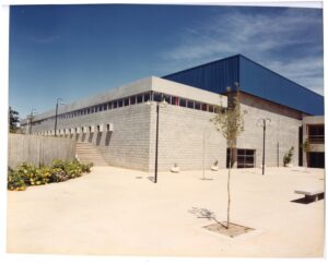 Vista externa do edifício, destacando a fachada em bloco de concreto e a cobertura metálica azul, com pátio, jardineiras e mobiliário urbano em primeiro plano — P0060 — SESI — Marília