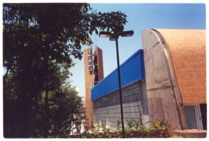 Vista externa do edifício do SESC, com fachada curva de tijolos, marquise azul e placa de identificação, emolduradas por árvores — P0131 — SESC - Piracicaba