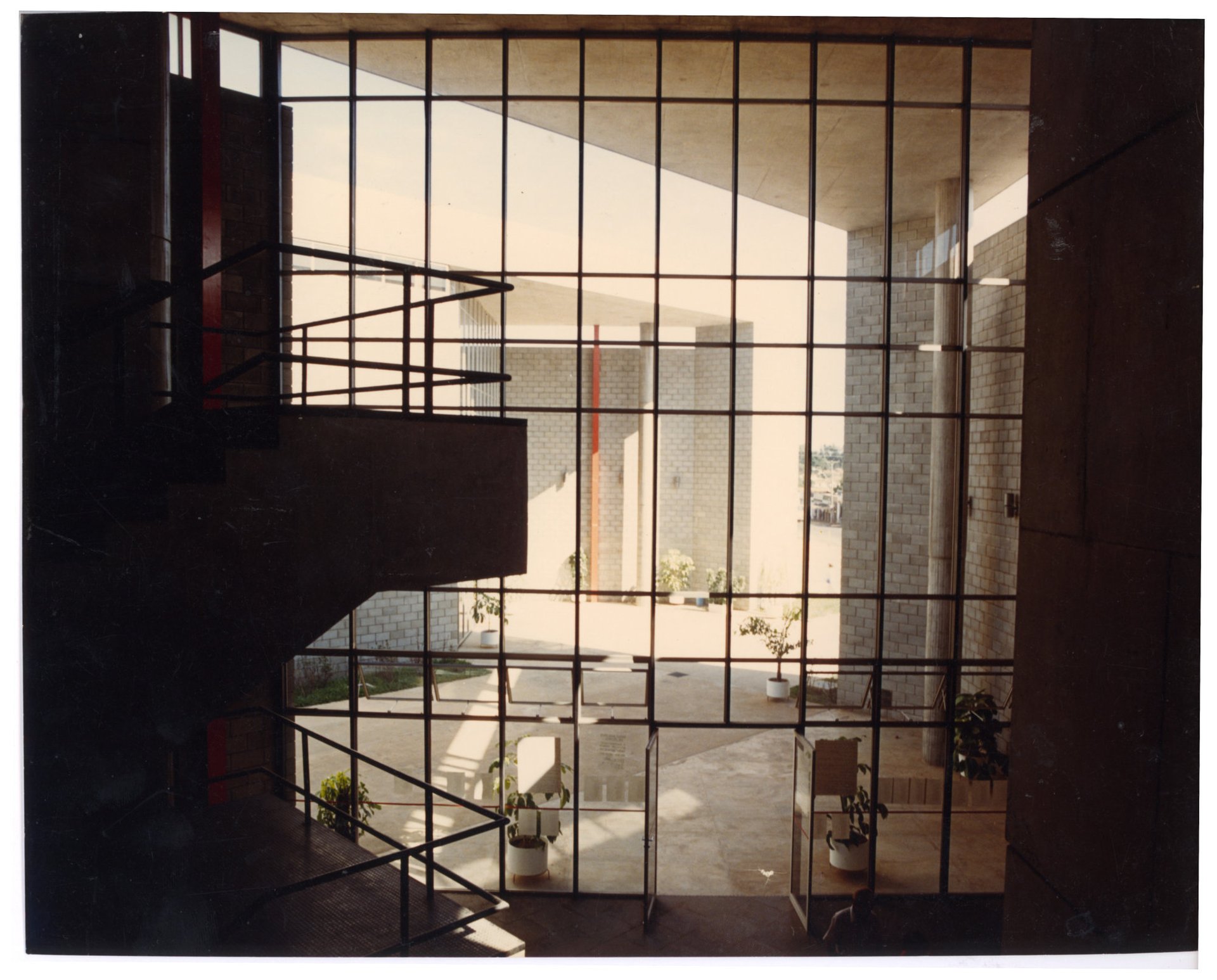 Vista interna a partir de escada, através de painel envidraçado quadriculado, para pátio externo com pilares revestidos e vasos de plantas — P0060 — SESI — Marília — F009-P0060-1984-S03-D00033