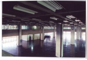 Vista interna de amplo pavimento desocupado, com pilares de concreto, piso escuro reflexivo e luminárias fluorescentes no teto — P0018 — Correios — Sede Regional e Centro de Triagem