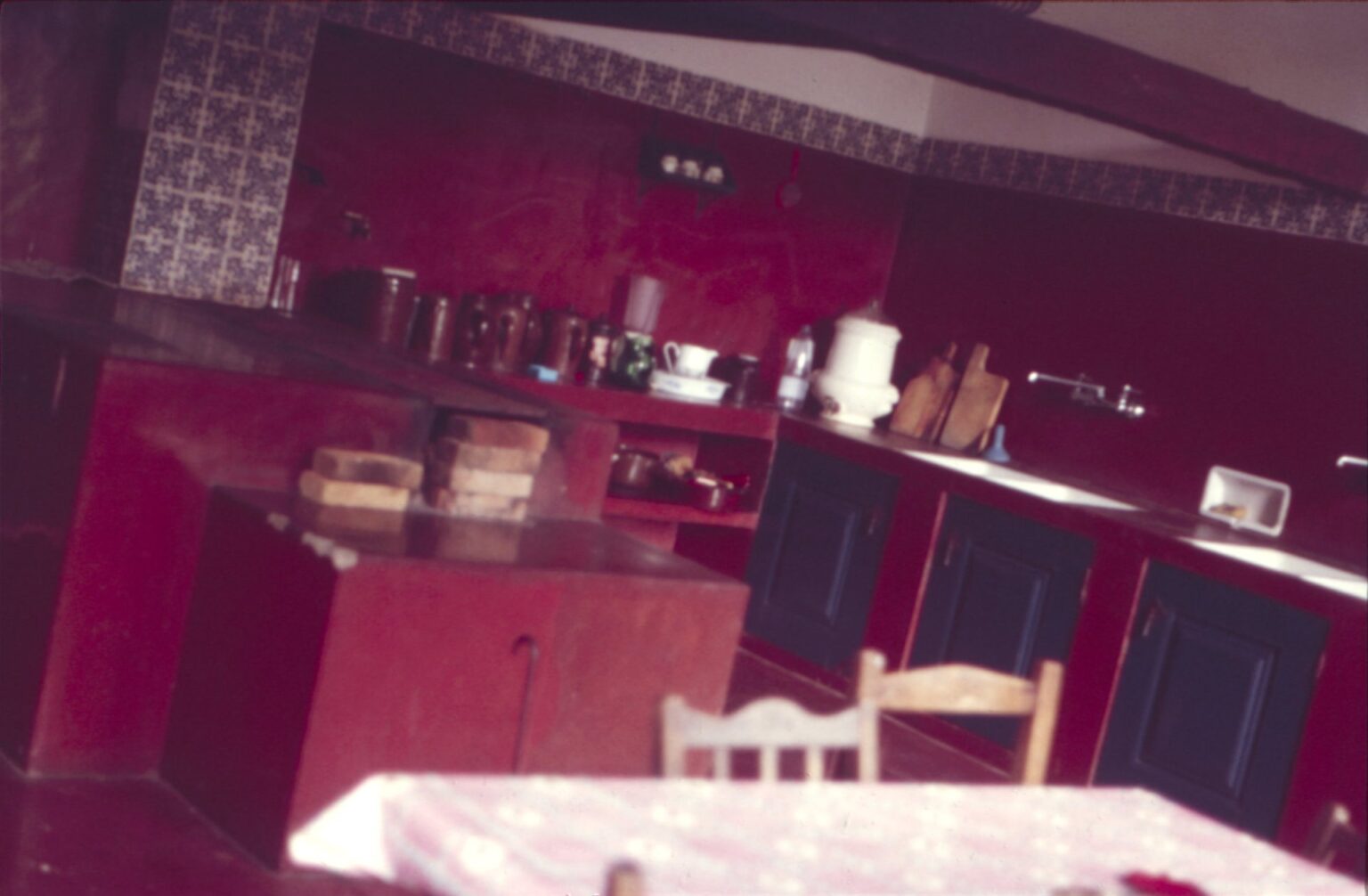 Vista interna de cozinha com armários vermelhos e azuis, utensílios sobre bancada e mesa em primeiro plano — P0024 — Fazenda Fernando Millan — F016-P0024-1981-S03-D00022