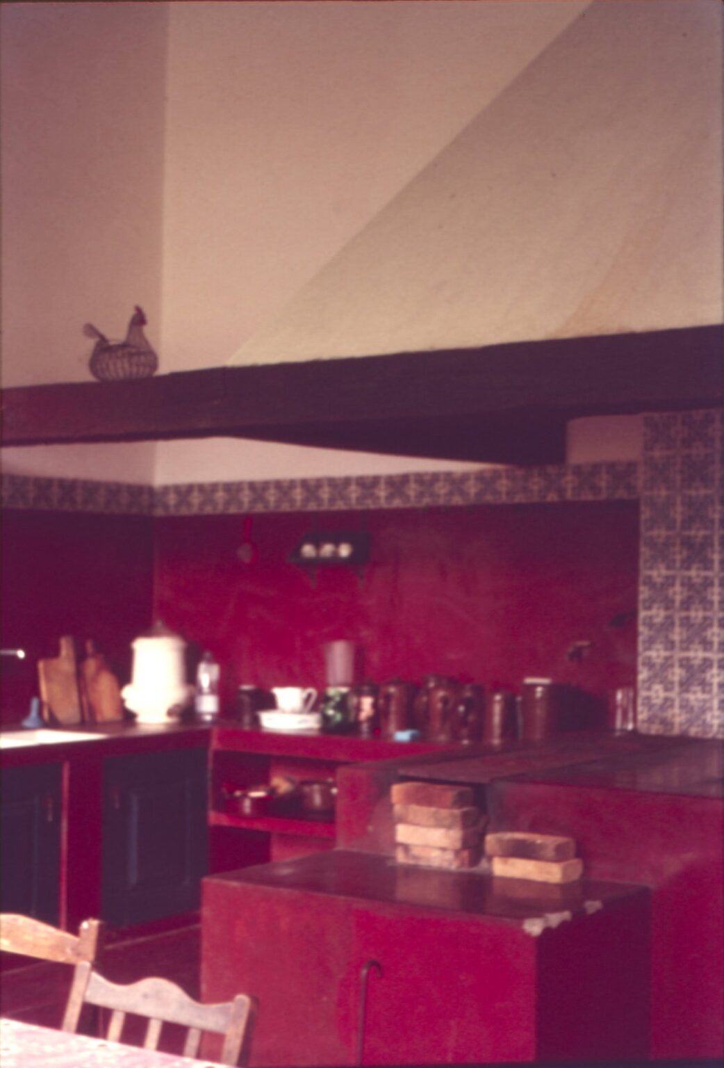 Vista interna de cozinha tradicional com bancadas de alvenaria pintadas de vermelho, azulejos decorativos e viga de madeira no teto — P0024 — Fazenda Fernando Millan — F016-P0024-1981-S03-D00012