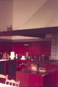 Vista interna de cozinha tradicional com bancadas de alvenaria pintadas de vermelho, azulejos decorativos e viga de madeira no teto — P0024 — Fazenda Fernando Millan