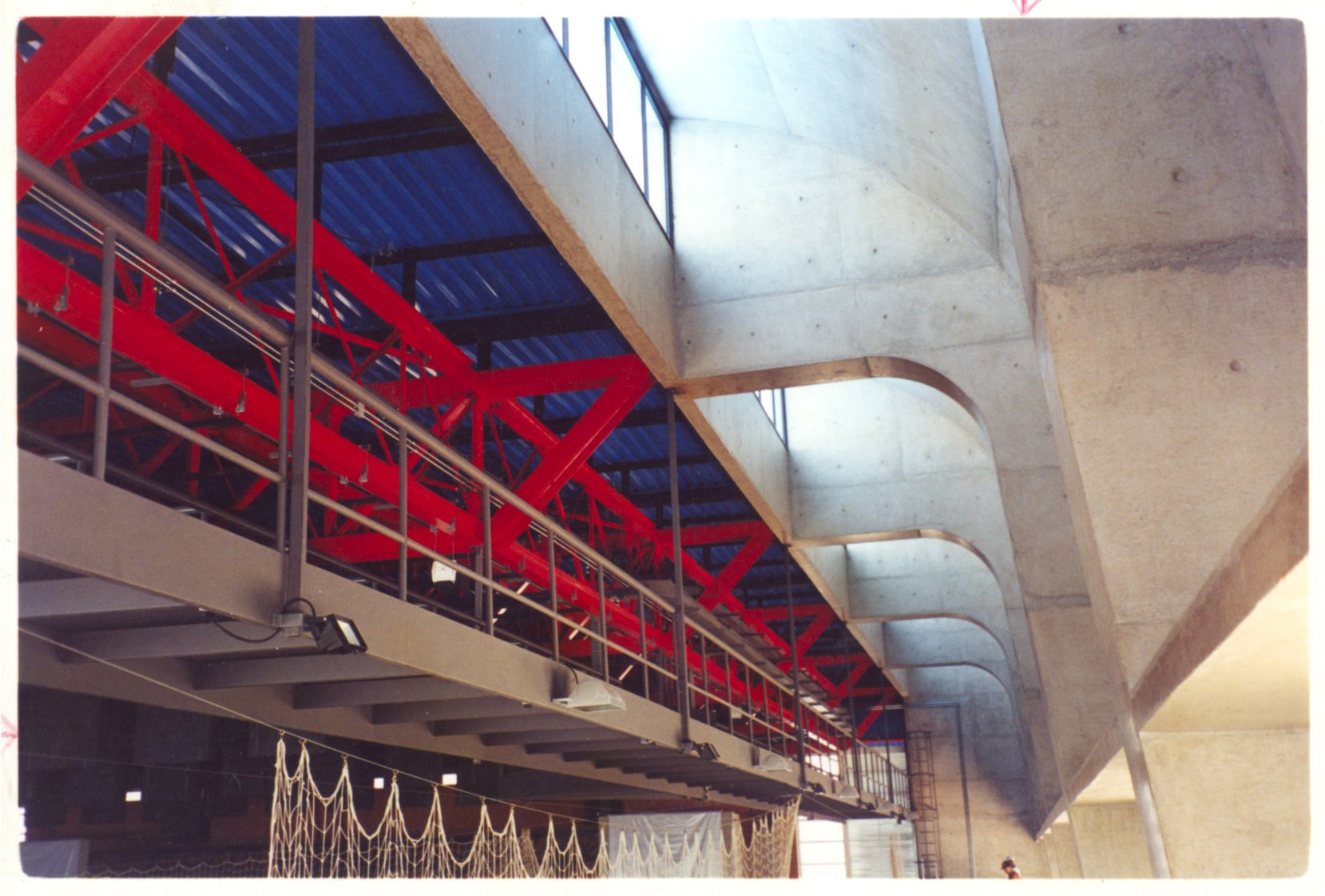 Vista interna de edifício em construção, destacando estrutura metálica vermelha treliçada sob cobertura de chapas azuis, ladeada por elementos curvos de concreto aparente — P0131 — SESC - Piracicaba — F009-P0131-0000-S03-D00002