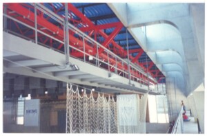 Vista interna de edifício mostrando estrutura de treliças metálicas vermelhas, pilares de concreto e cobertura têxtil em formato ondulado, com faixa escrita "SESC" ao fundo — P0131 — SESC - Piracicaba