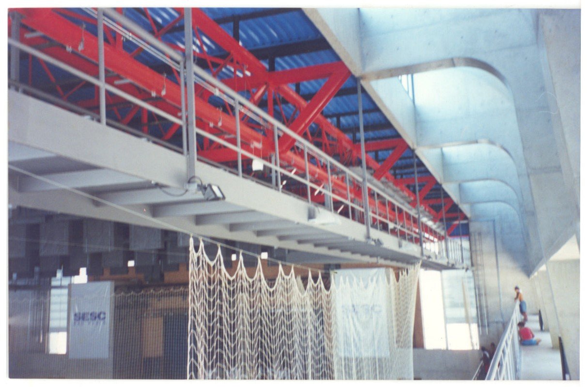 Vista interna de edifício mostrando estrutura de treliças metálicas vermelhas, pilares de concreto e cobertura têxtil em formato ondulado, com faixa escrita "SESC" ao fundo — P0131 — SESC - Piracicaba — F009-P0131-0000-S03-D00044