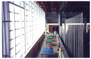 Vista interna de ginásio esportivo com mesas de tênis de mesa, cobertura em painéis translúcidos e rede de proteção suspensa — P0131