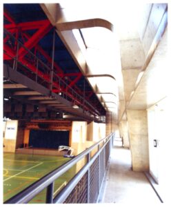 Vista interna de ginásio esportivo, mostrando estrutura metálica e elementos de concreto, com quadra ao fundo e placas identificadas como 'SESC — P0131