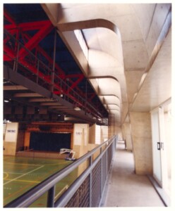 Vista interna de ginásio esportivo, mostrando estrutura metálica e elementos de concreto, com quadra ao fundo e placas identificadas como 'SESC — P0131