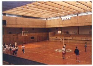 Vista interna de ginásio poliesportivo mostrando quadra esportiva com piso de madeira, tabela de basquete, rede de vôlei, arquibancadas e grupo de pessoas na quadra — P0015 — SESI — Ribeirão Preto