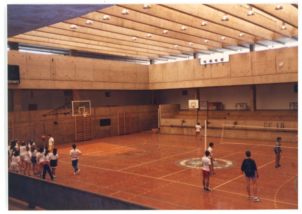 Vista interna de ginásio poliesportivo mostrando quadra esportiva com piso de madeira, tabela de basquete, rede de vôlei, arquibancadas e grupo de pessoas na quadra — P0015 — SESI — Ribeirão Preto — F009-P0015-1973-S03-D00057