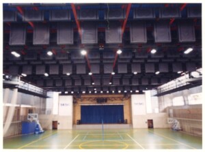 Vista interna de quadra poliesportiva coberta, com estrutura metálica aparente no teto, palco com cortina azul ao fundo e piso de quadra demarcado — P0131 — SESC - Piracicaba