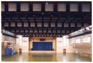 Vista interna de quadra poliesportiva coberta, com palco de cortina azul ao fundo e banners "SESC" nas laterais — P0131 — SESC - Piracicaba
