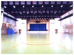 Vista interna de quadra poliesportiva coberta, com piso demarcado para diversas modalidades e palco com cortina azul ao fundo — P0131