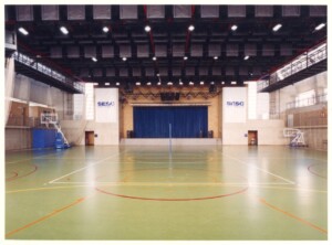 Vista interna de quadra poliesportiva coberta, com piso demarcado para diversas modalidades e palco com cortina azul ao fundo — P0131 — SESC - Piracicaba