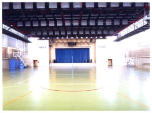 Vista interna de quadra poliesportiva do SESC, com piso demarcado para esportes e palco com cortina azul ao fundo — P0131