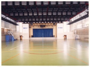 Vista interna de quadra poliesportiva do SESC, com piso demarcado para esportes e palco com cortina azul ao fundo — P0131 — SESC - Piracicaba