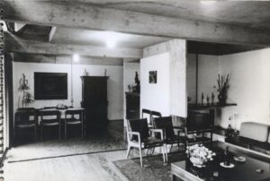 Vista interna de sala de estar e jantar integradas, com teto de vigas de concreto aparente, mobiliário diverso e objetos decorativos — P0001 — Residência Teresa Martino